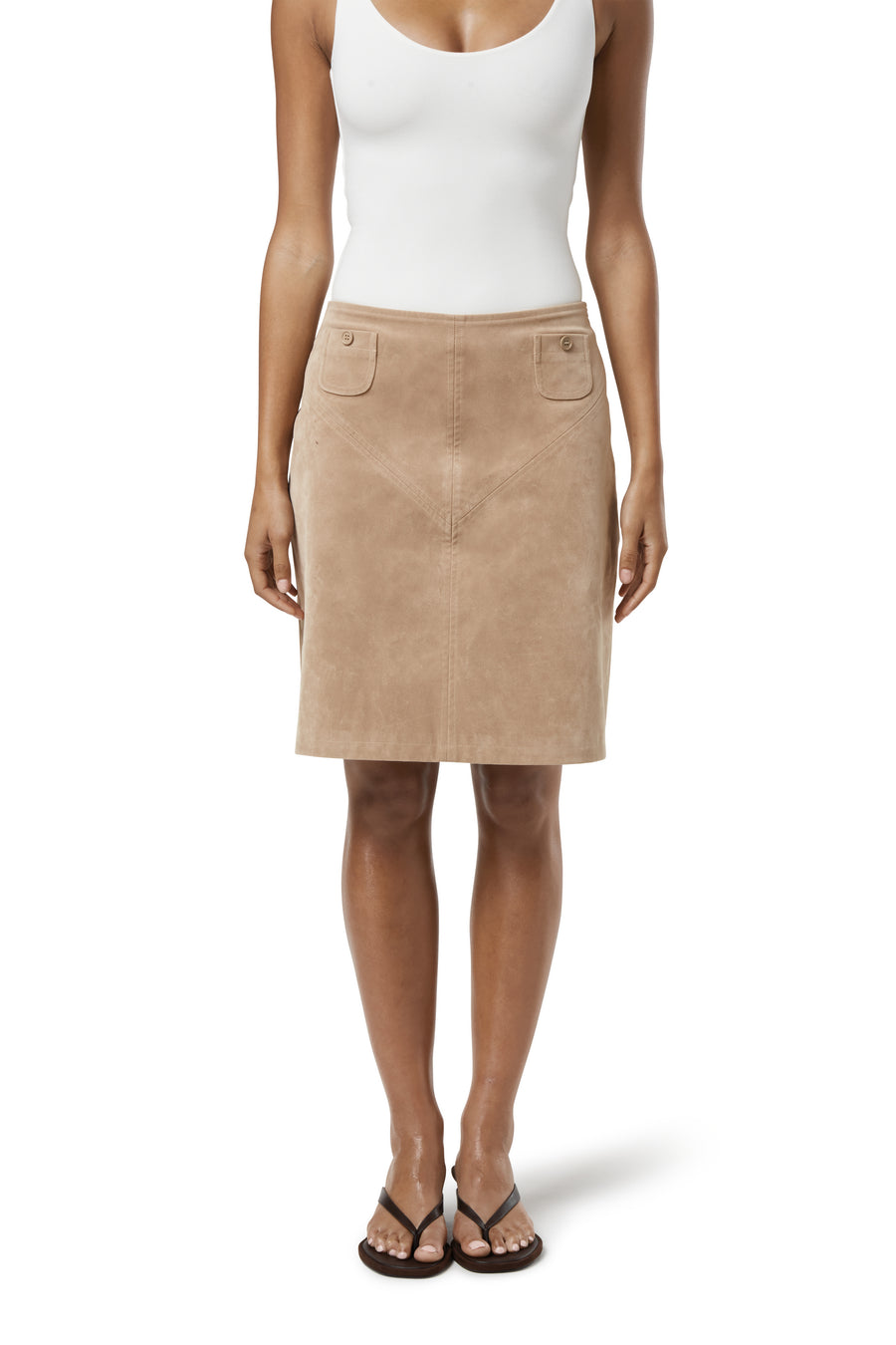 Woman wearing a fitted beige suede knee-length skirt with front flap pockets, paired with a white tank top, black flip-flop sandals, and a shell necklace.